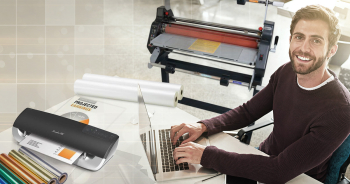 Office employee using a computer next to laminating machines and supplies at his desk.